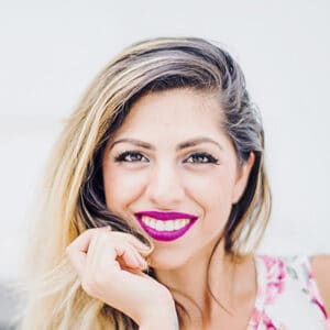A smiling person with long, light hair and bright pink lipstick, resting their chin on their hand while wearing a floral top.